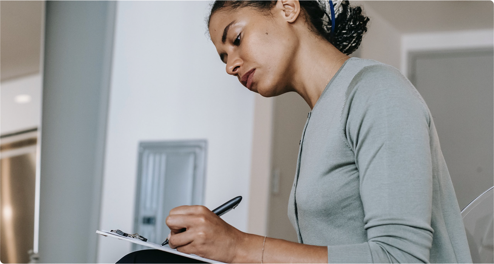 Woman writing on a clipboard while seated in a light-colored room.